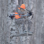 Hunter in camo perched on a metal treestand in a snowy forest, wearing a bright orange hat and vest and aiming a crossbow.