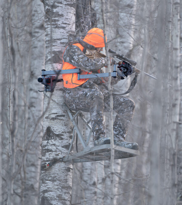 Hunter in camo perched on a metal treestand in a snowy forest, wearing a bright orange hat and vest and aiming a crossbow.