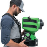 Man in profile wearing a cap and harness, holding a green rectangular industrial device with a control panel and dials on the side.